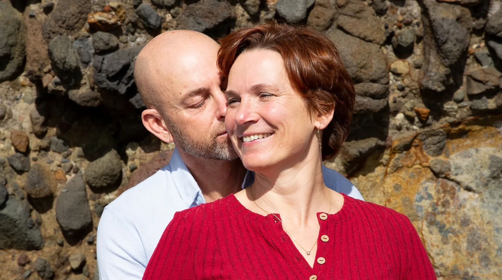 Mark holding Tarisha from behind, eyes closed, on a rocky beach at Piha
