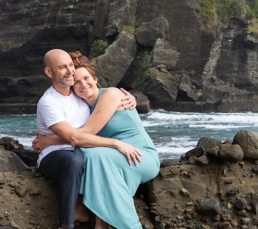 Tarisha and Mark hugging on the rocks by the sea at Piha